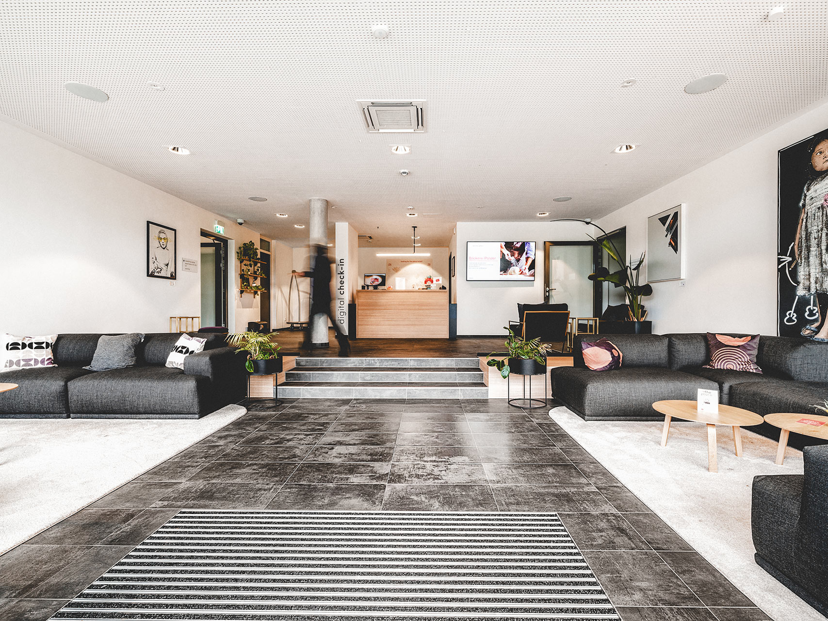 Weitläufige Lobby der Serviced Apartments in Herzogenaurach mit dunklen Sofalandschaften, digitalem Check-in-Tresen und dunklem Fliesenboden